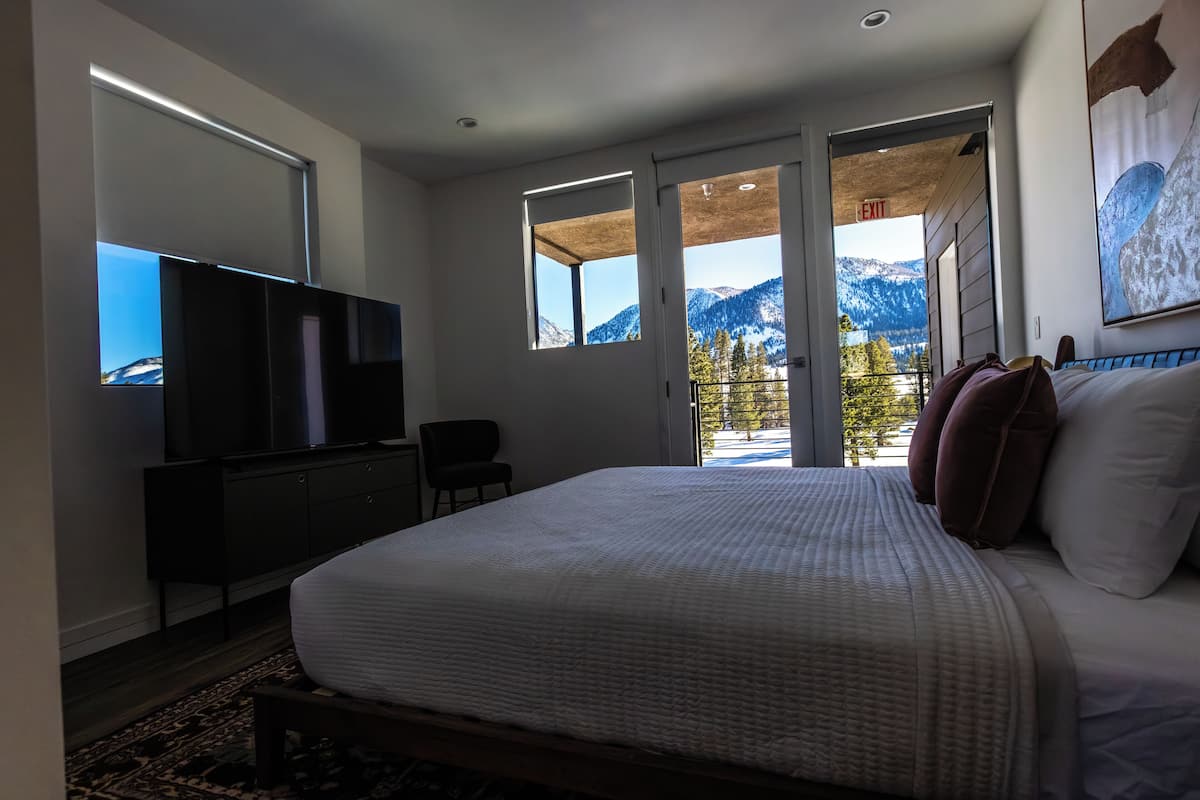 A bedroom with a large bed, a TV, a desk, and a chair. There are large windows with a mountain view.