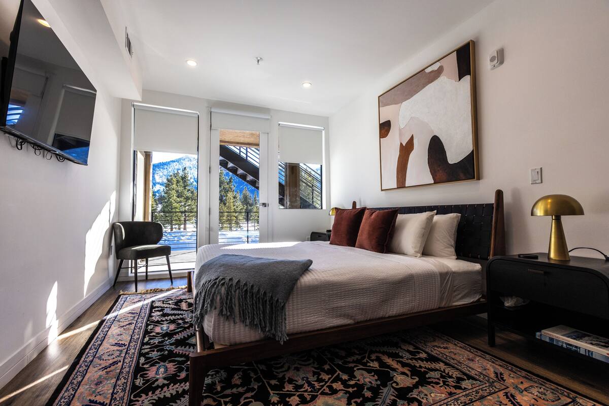 A modern bedroom with a large bed, a TV mounted on the wall, and a view of a mountain through a sliding door.