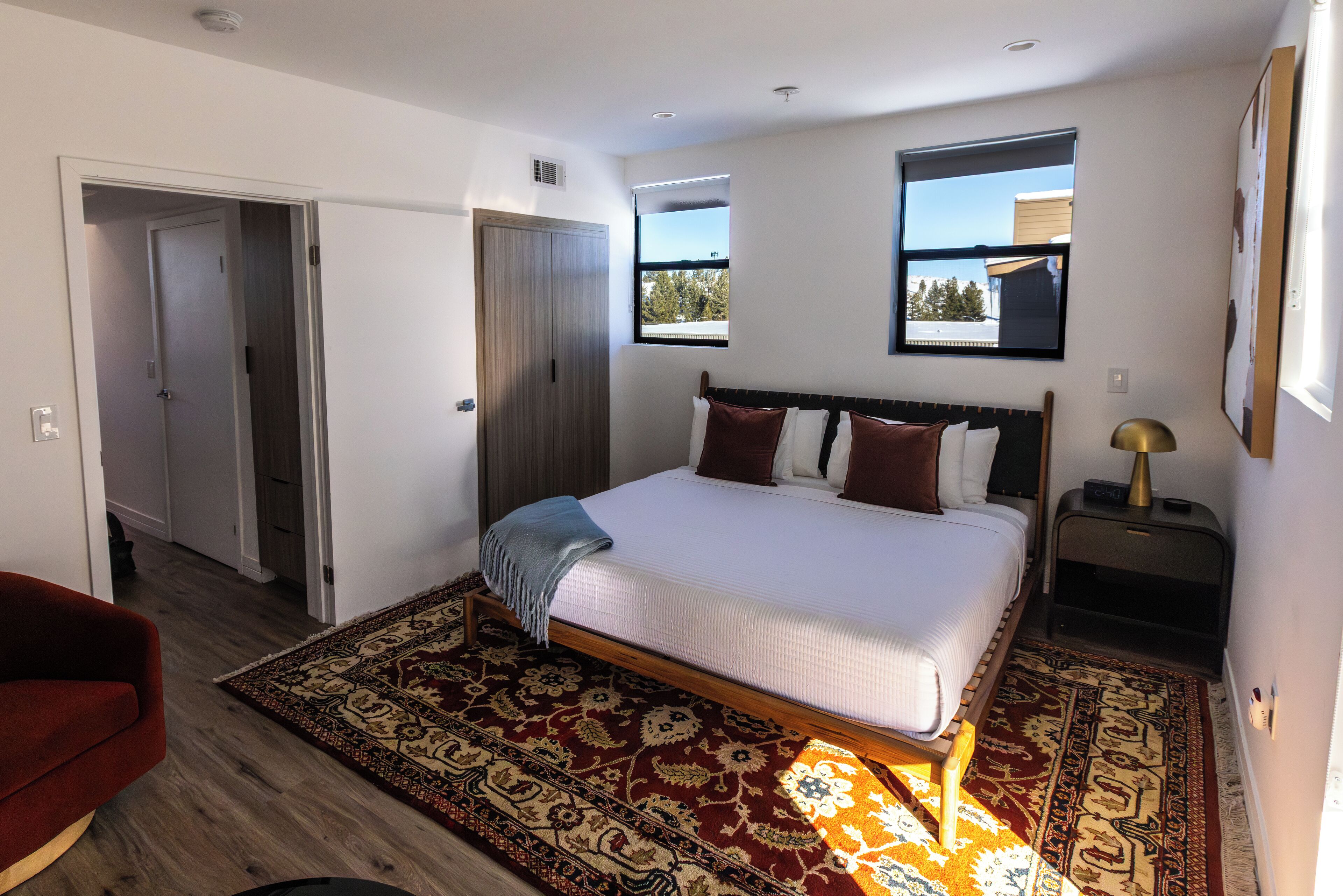 A bedroom with a large bed, a red chair, a wooden floor, a patterned rug, and a window with a view of buildings.