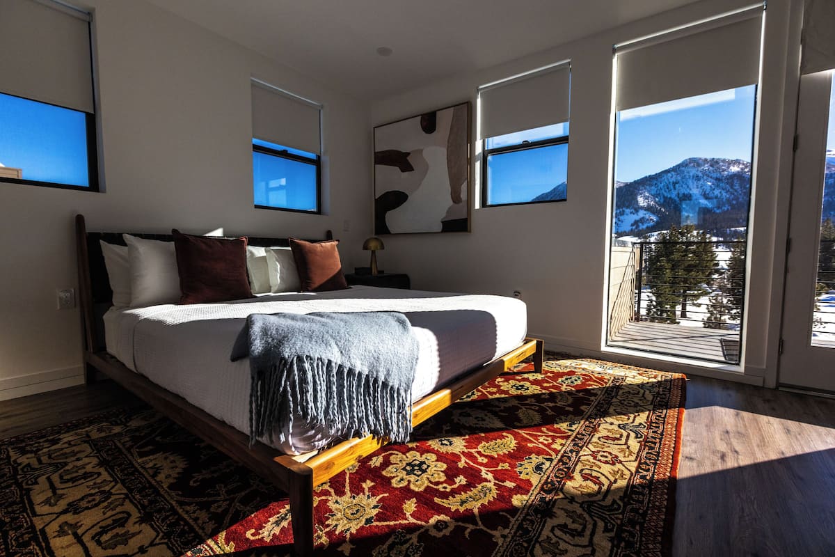 A bedroom with a large bed, a red and beige patterned rug, and a mountain view through the window.