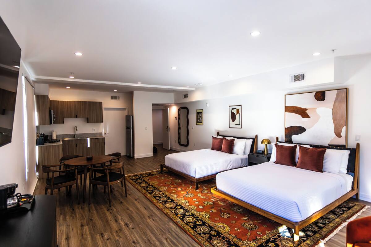 A modern hotel room with two beds, a dining area, and a kitchenette.