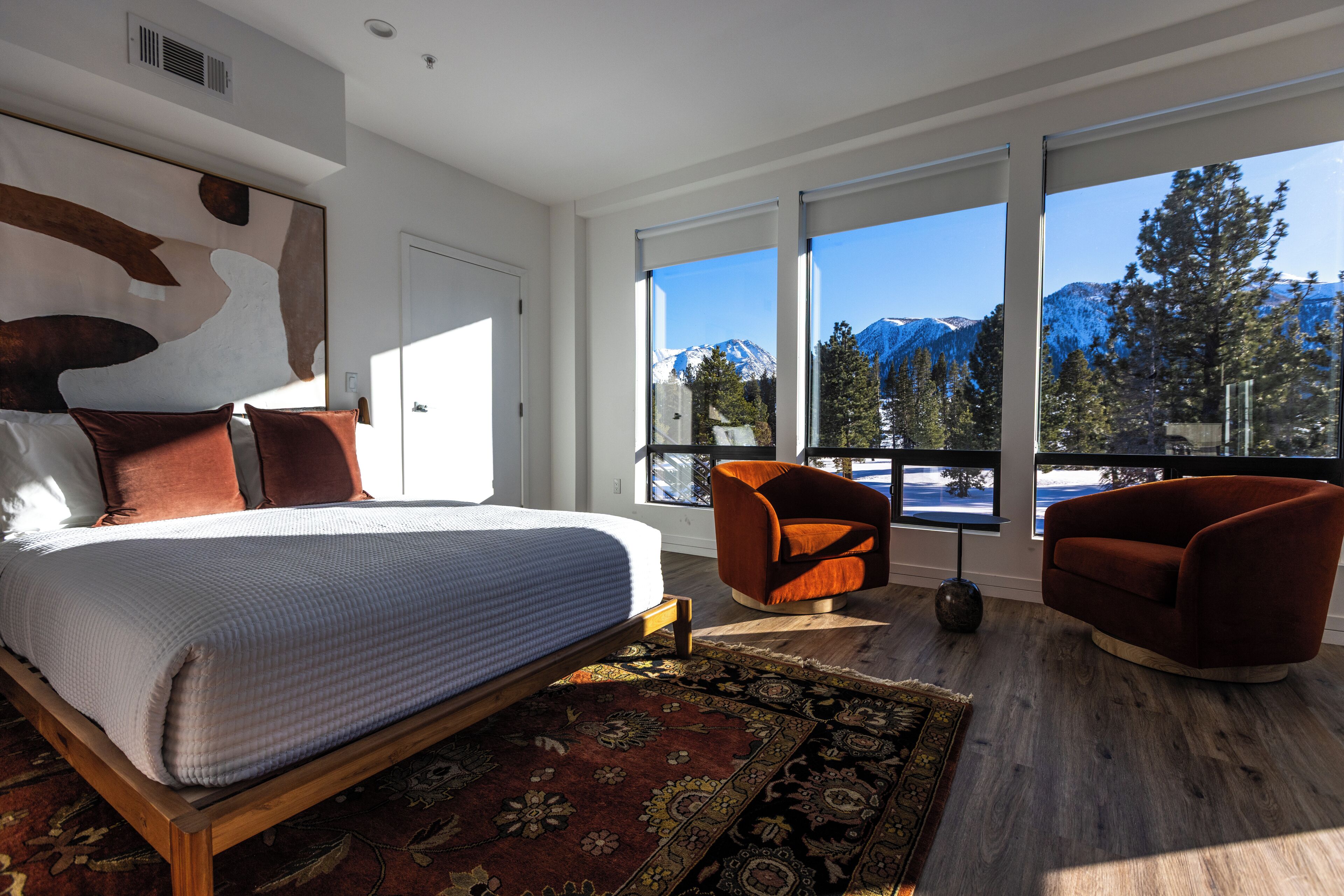 A modern bedroom with a large bed, two armchairs, and a view of snow-capped mountains through large windows.