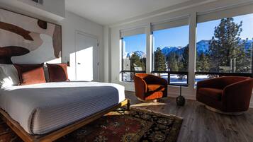 A modern bedroom with a large bed, two armchairs, and a view of snow-capped mountains through large windows.