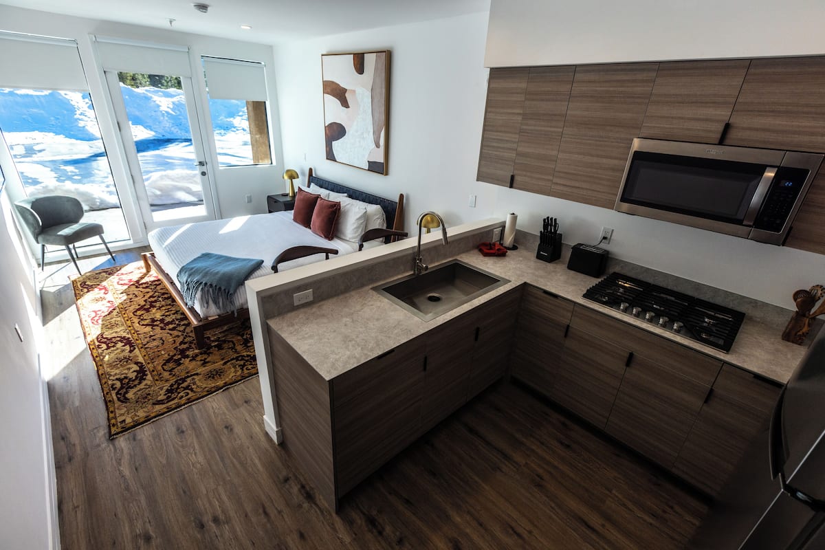 A modern hotel room with a large bed, a built-in kitchen with a microwave, and a view of a snowy landscape through the sliding doors.