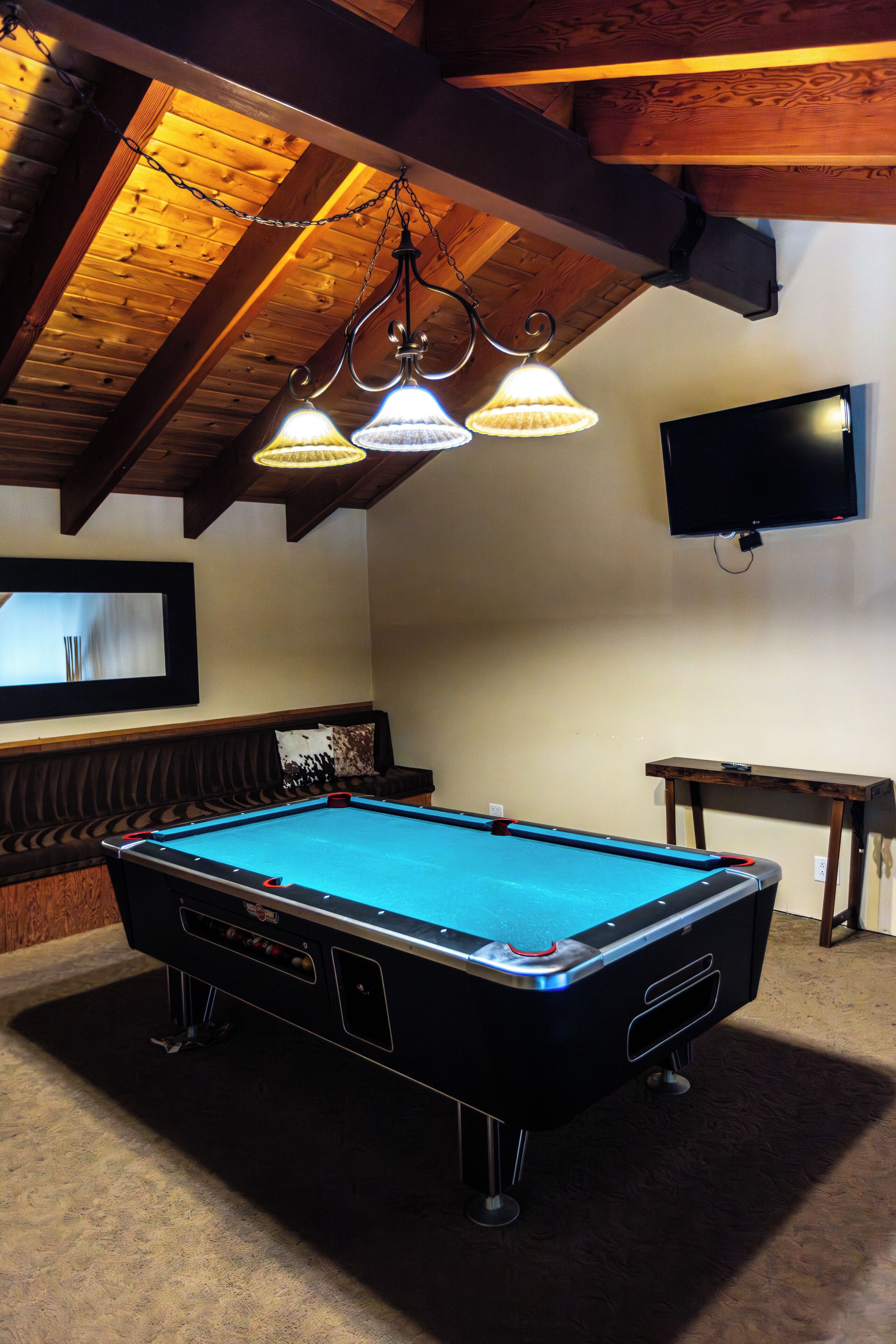 Una mesa de billar con superficie de fieltro azul, un sofá de cuero marrón, un televisor montado en la pared y un techo de madera con vigas a la vista.