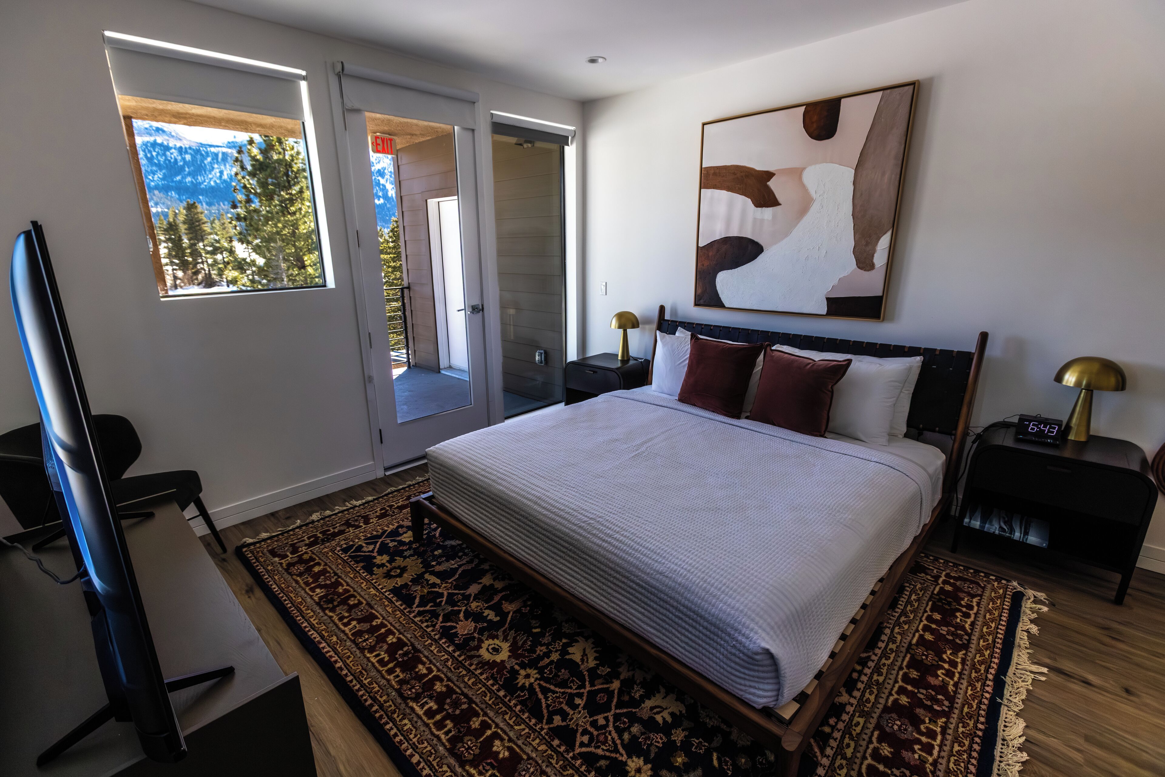 A bedroom with a large bed, a TV, a desk, and a window with a view of trees and mountains.