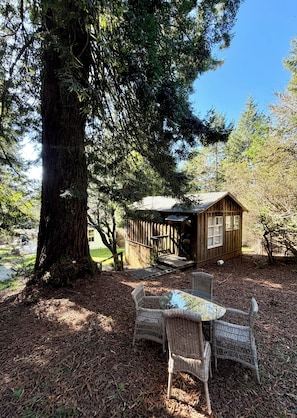 A wooden cabin with a porch, surrounded by trees and a glass table with wicker chairs.