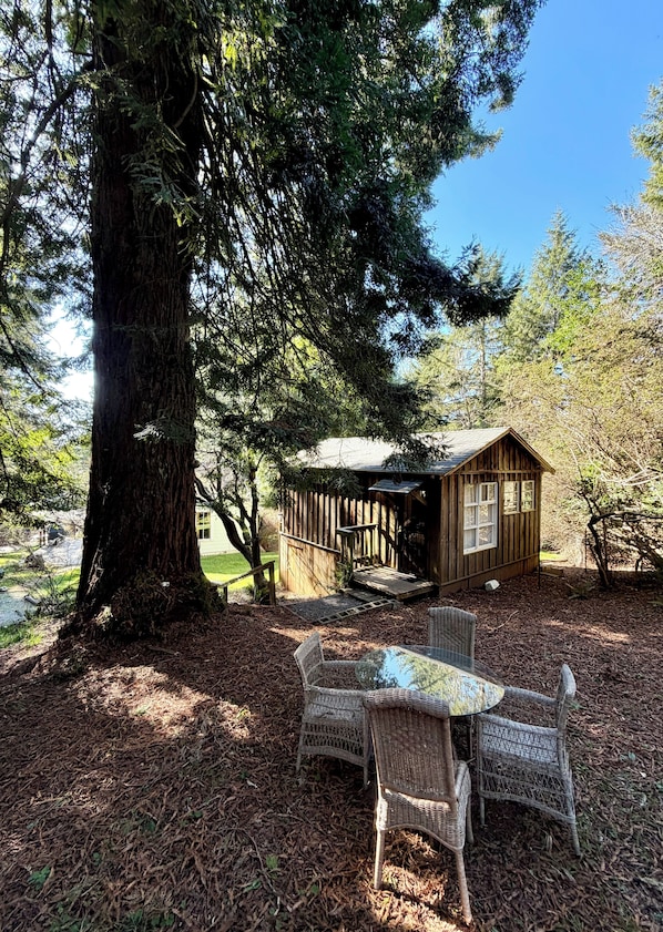 Une cabane en bois avec un porche, entourĂ©e dâarbres, et une table en verre avec des chaises en osier.