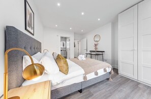 A modern bedroom with a grey upholstered headboard, a bedside table with a lamp, a bed with white and gold bedding, a mirror, and a wardrobe. - 3 bedroom cozy flat (London)
