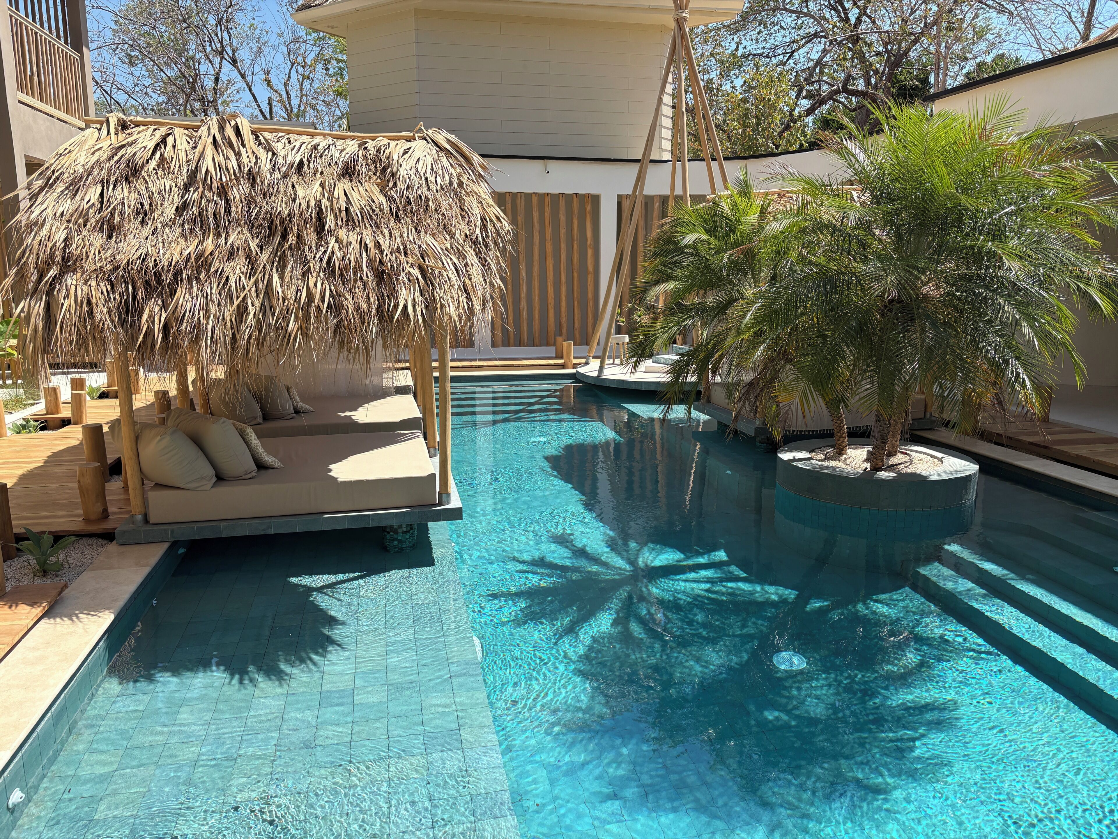 Une piscine dotée d’un espace détente avec toit de chaume et de palmiers.