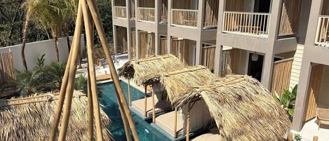 A poolside area with thatched umbrellas and lounge chairs, a clear pool, and a multi-story building with balconies.