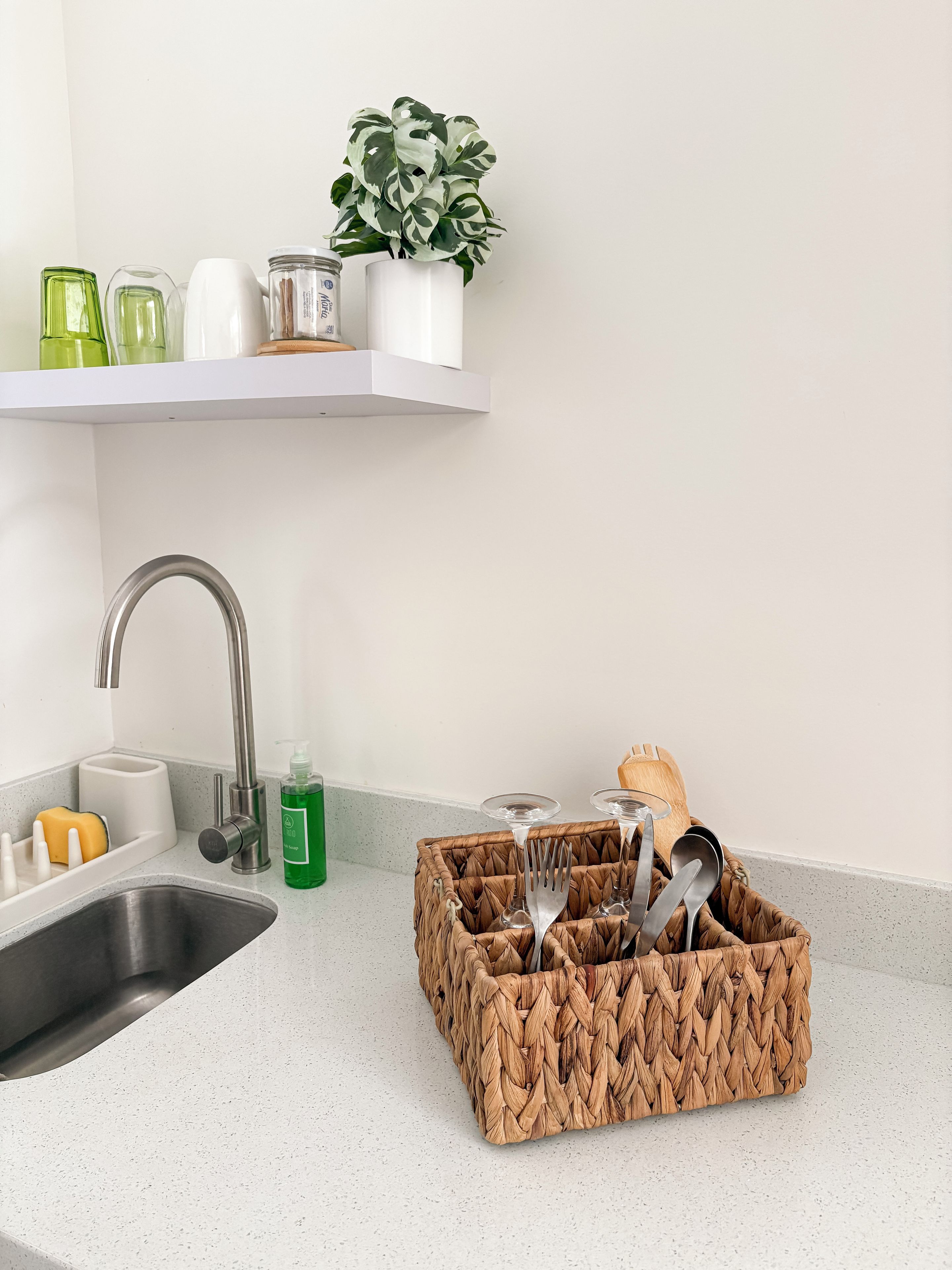 A kitchen with a stainless steel sink, a white countertop, a hanging plant, and a woven basket containing utensils.