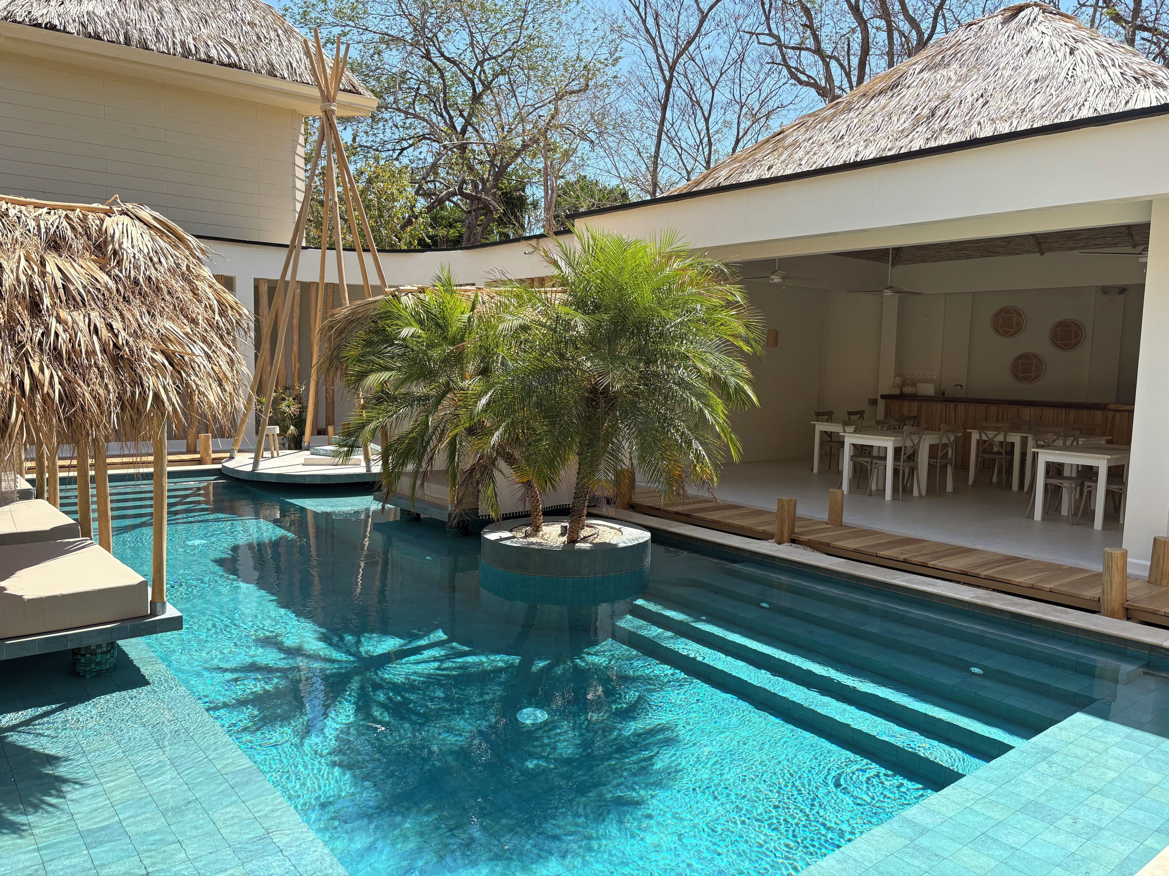 A swimming pool with a thatched roof structure, palm trees, and a dining area with white furniture.