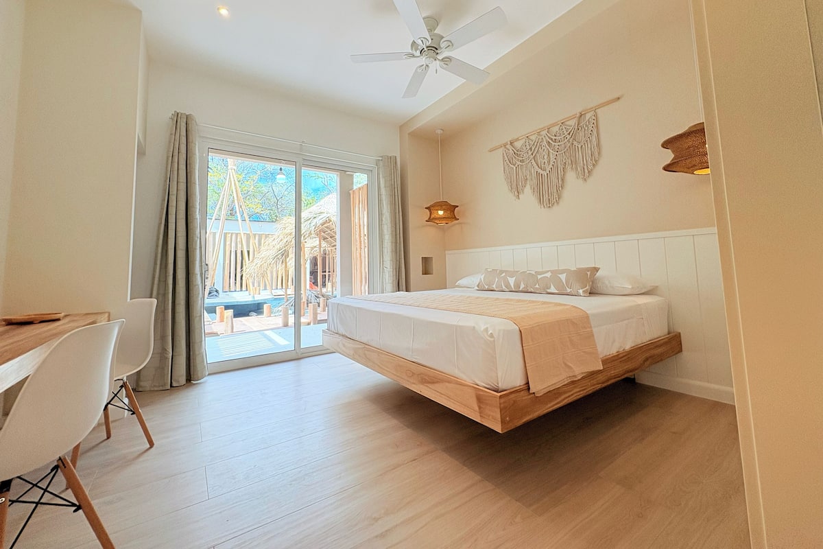 A modern bedroom with a bed, a desk, and a chair. There is a ceiling fan and a macrame wall hanging.