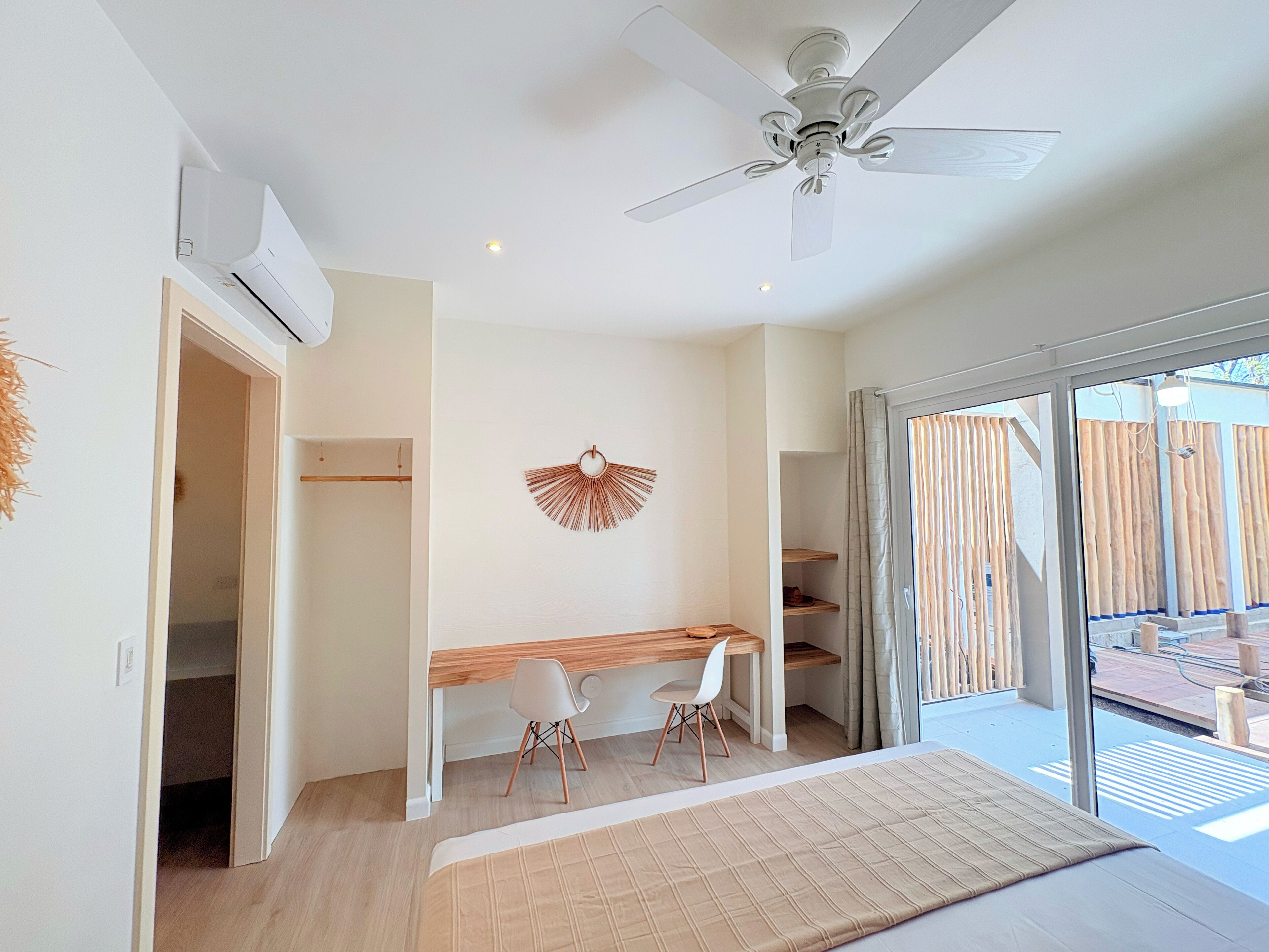 A modern bedroom with a bed, a desk, two chairs, a ceiling fan, and a view of the outside.