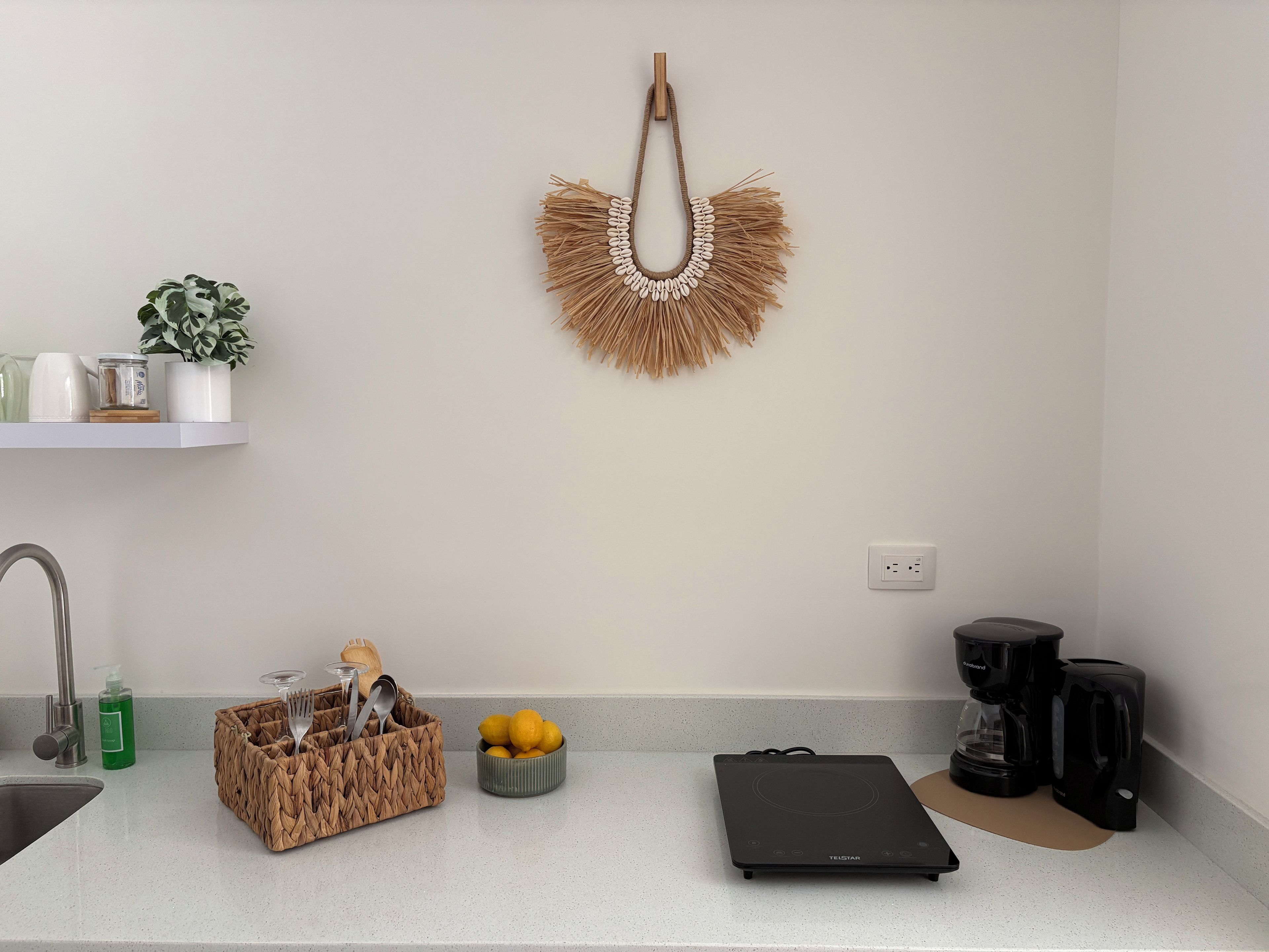 A kitchen with a countertop, a sink, a coffee maker, a kettle, a fruit bowl, a basket with utensils, and a wall-mounted decorative item.