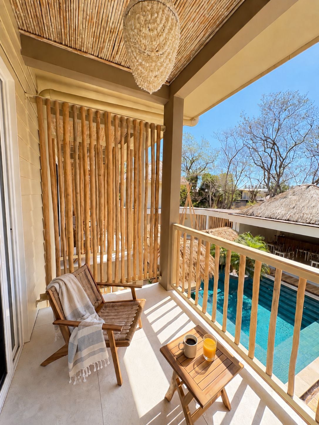 Un balcone con vista su una piscina, una sedia in vimini, un tavolo con un drink e una lampada decorativa a soffitto.