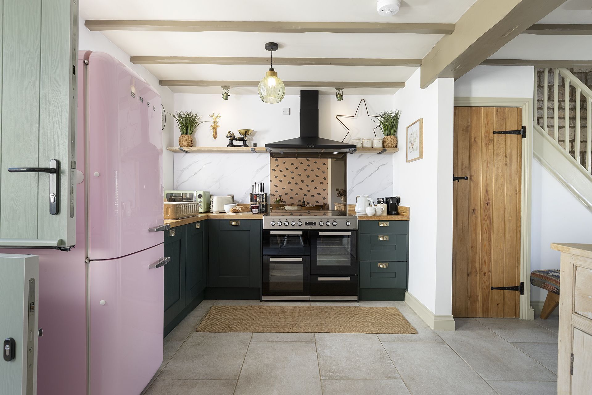 Una cuina amb armaris verds fosc, un frigorífic rosa, un forn integrat i una escala de fusta que condueix a una altra habitació.