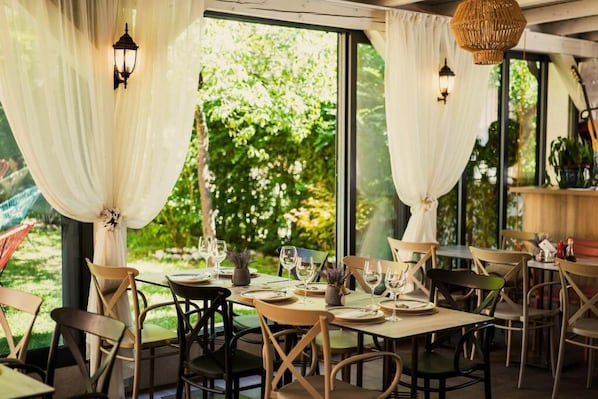 Un área de comedor al aire libre con mesas preparadas para dos, rodeada de sillas y con cortinas blancas.