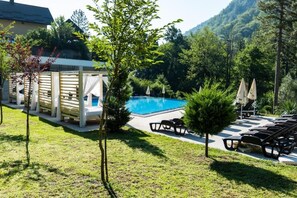 Un área de piscina con sillones de descanso, una estructura de madera con cortinas blancas y follaje circundante.