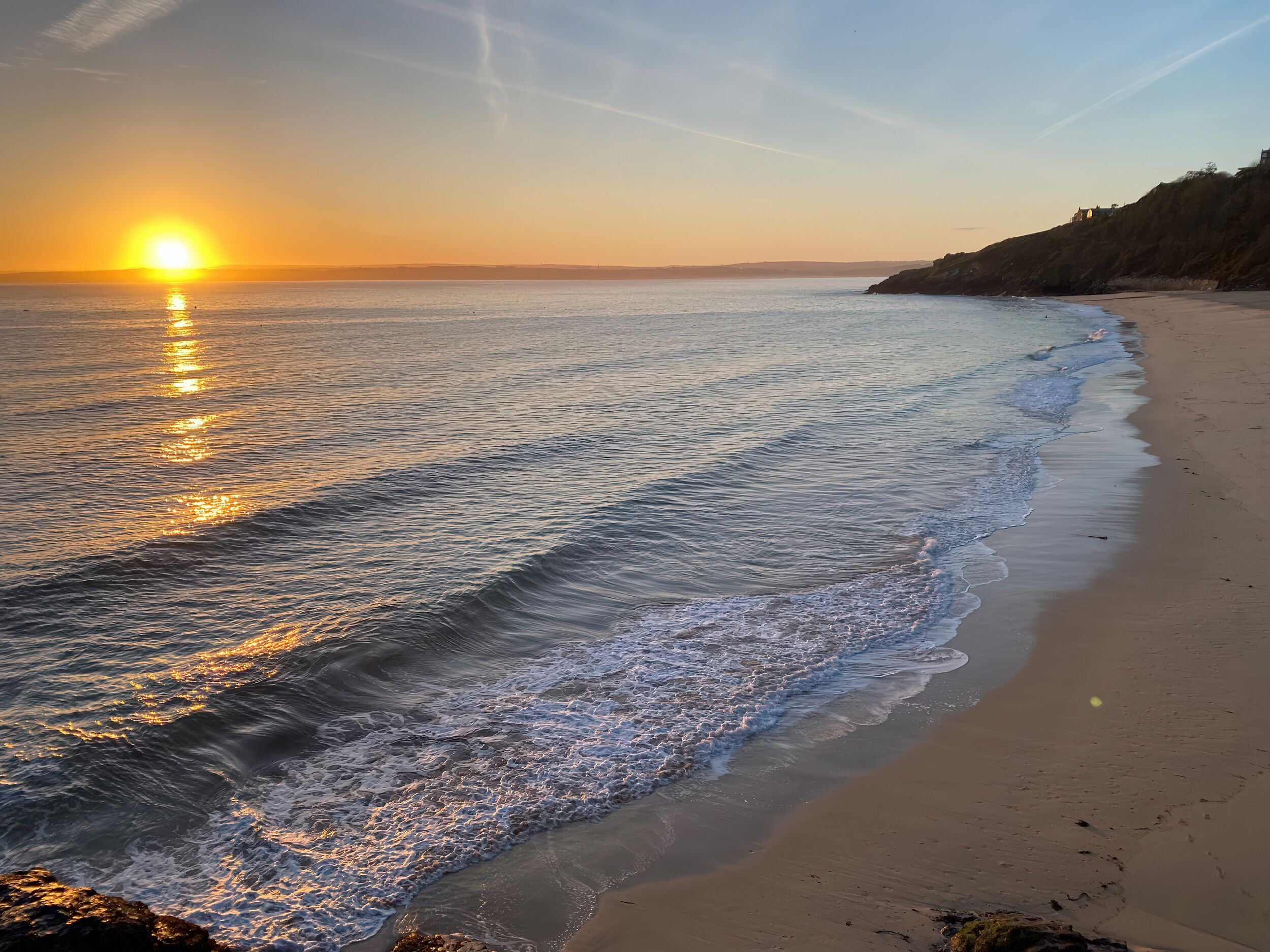 Ein Strand mit Sonnenuntergang, ruhiger Wellengang und einer felsigen Küstenlinie.