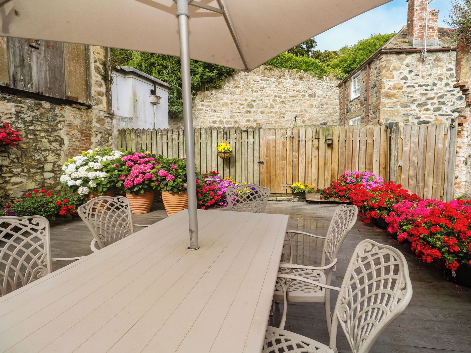 A patio with a table and chairs, surrounded by stone walls and wooden fences, with potted plants and flowers.