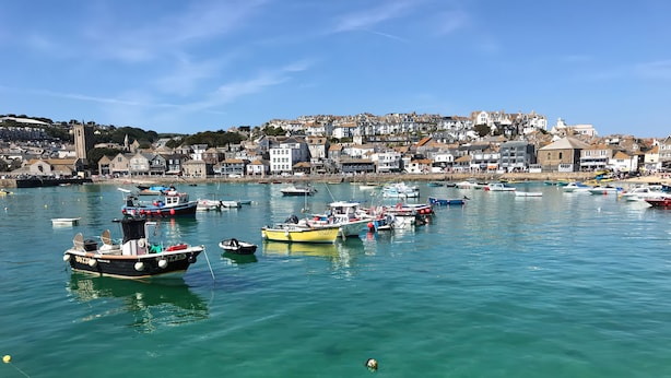 A harbor with various boats docked, clear water, and a town in the background. - Ammil Bespoke (St Ives)