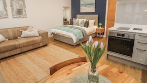 A modern bedroom with a bed, a sofa, a wooden table with a vase, and a kitchen area with an oven. - Ammil Bespoke (St Ives)