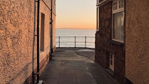 A narrow pathway leading to a waterfront view, flanked by stone buildings with windows and a metal railing. - Ammil Bespoke (St Ives)