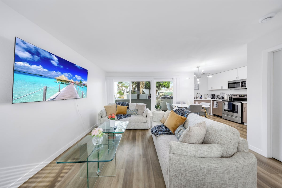 Un salon moderne avec une table basse en verre, un canapé beige et un grand tableau représentant une scène de plage tropicale.