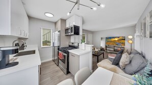 Een moderne keuken met een inbouwmagnetron, fornuis en spoelbak. Er is een eethoek met een tafel en stoelen, en een bank met kussens.