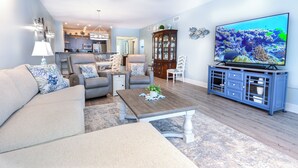Ein Wohnzimmer mit einer Couch, Sesseln, einem Couchtisch, einem Fernseher und einer Küche im Hintergrund.