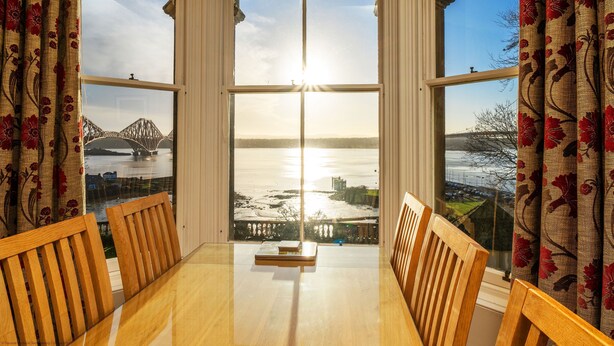Ein Zimmer mit großem Fenster mit Blick auf ein Gewässer und eine Brücke, Holzstühle und ein Tisch mit einem Buch.
