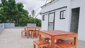 A rooftop patio with wooden tables and chairs, a white building with black-framed windows, and a view of greenery and palm trees in the background. - Crescent Bay Weligama (Weligama)