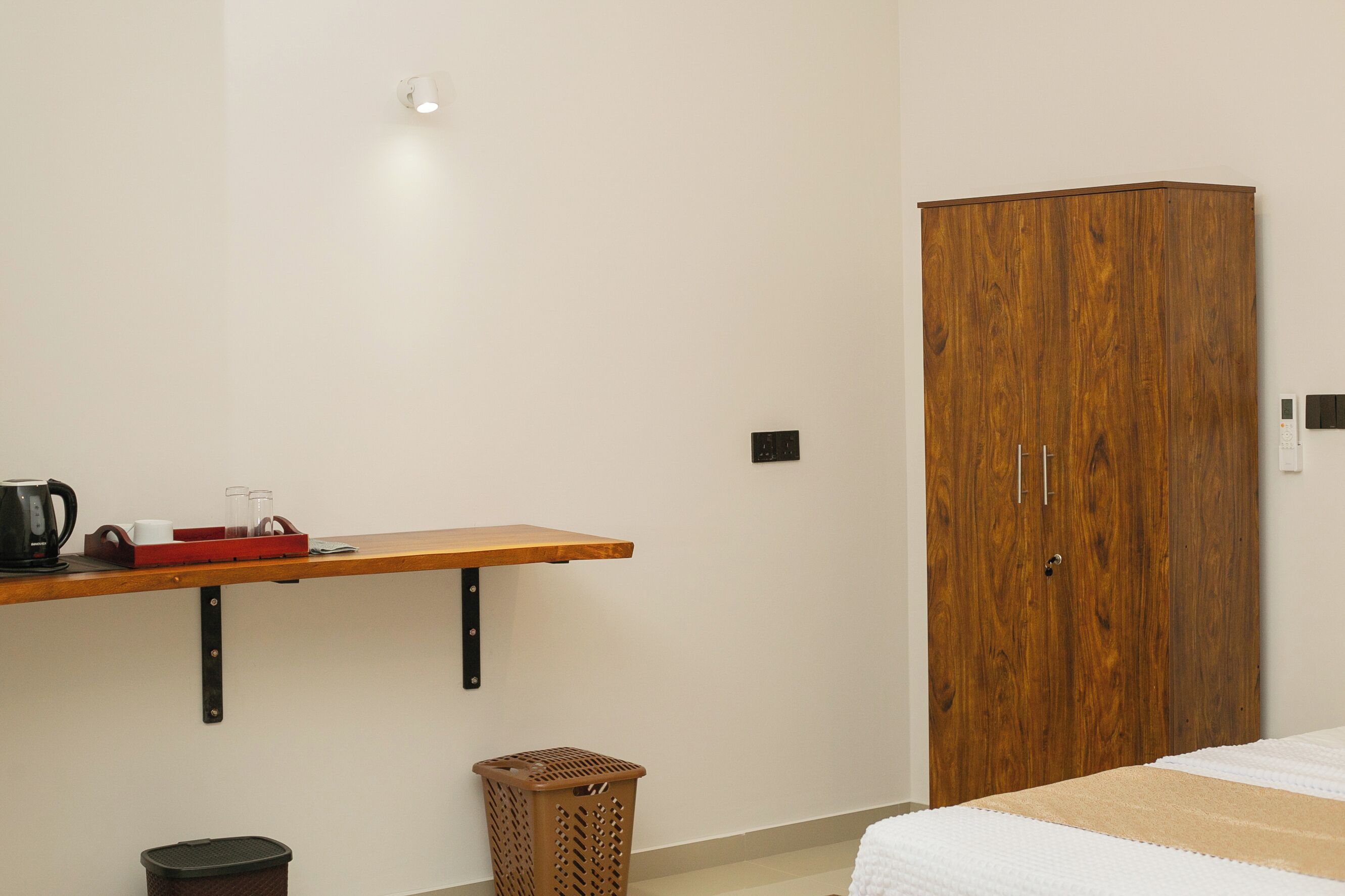 A bedroom with a wooden wardrobe, a wooden shelf with a coffee maker, glasses, and a red tray, a wicker basket, and a white bedspread.