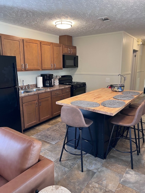 A kitchen with wooden cabinets, a black refrigerator, a microwave, a stove, and a dining area with a wooden table and two bar stools. - Pet Friendly Cozy 2-bedroom manufactured home with accessible feature's (North Fort Myers)