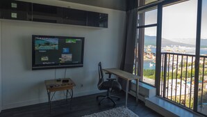 Una habitación moderna con un ventanal grande, un televisor montado, un escritorio y una silla.