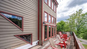 Eine Holzterrasse mit Adirondack-Stühlen, einem Holzgeländer und einem Haus mit mehreren Fenstern und einer Tür.