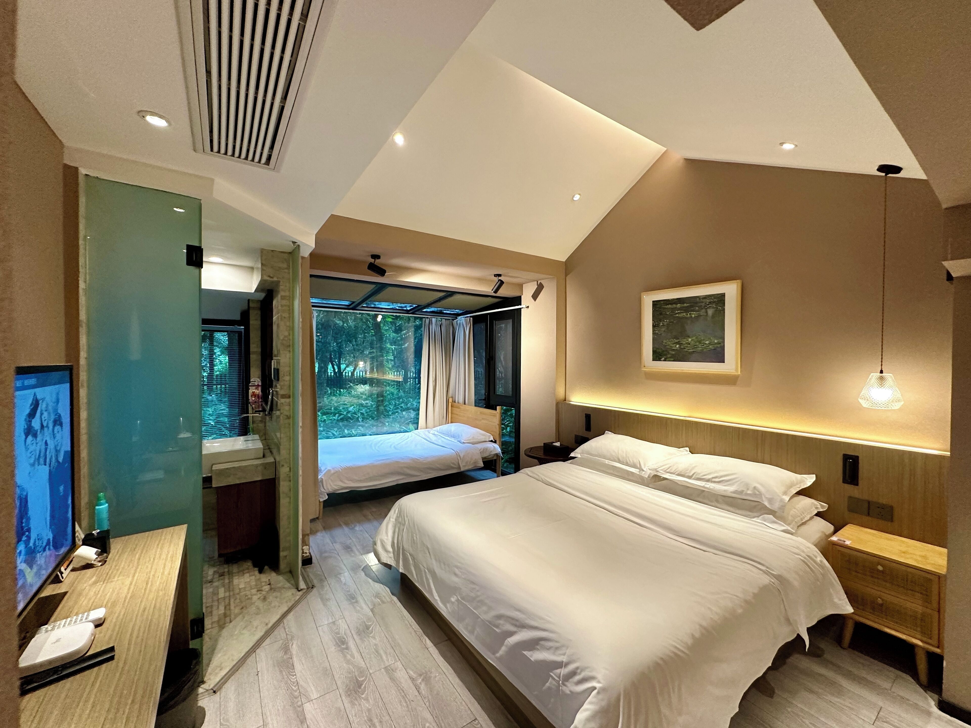 A modern hotel room with a large bed, a desk with a laptop, and a view of a forest through a glass door.