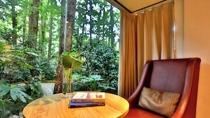 A wooden table with a book and a vase, a leather chair with a cushion, and a view of a lush garden through a window with curtains. - Lotus Glade Hotel (Hangzhou)