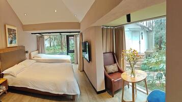 A hotel room with two beds, a TV, a small table with a vase, and a view of a garden.