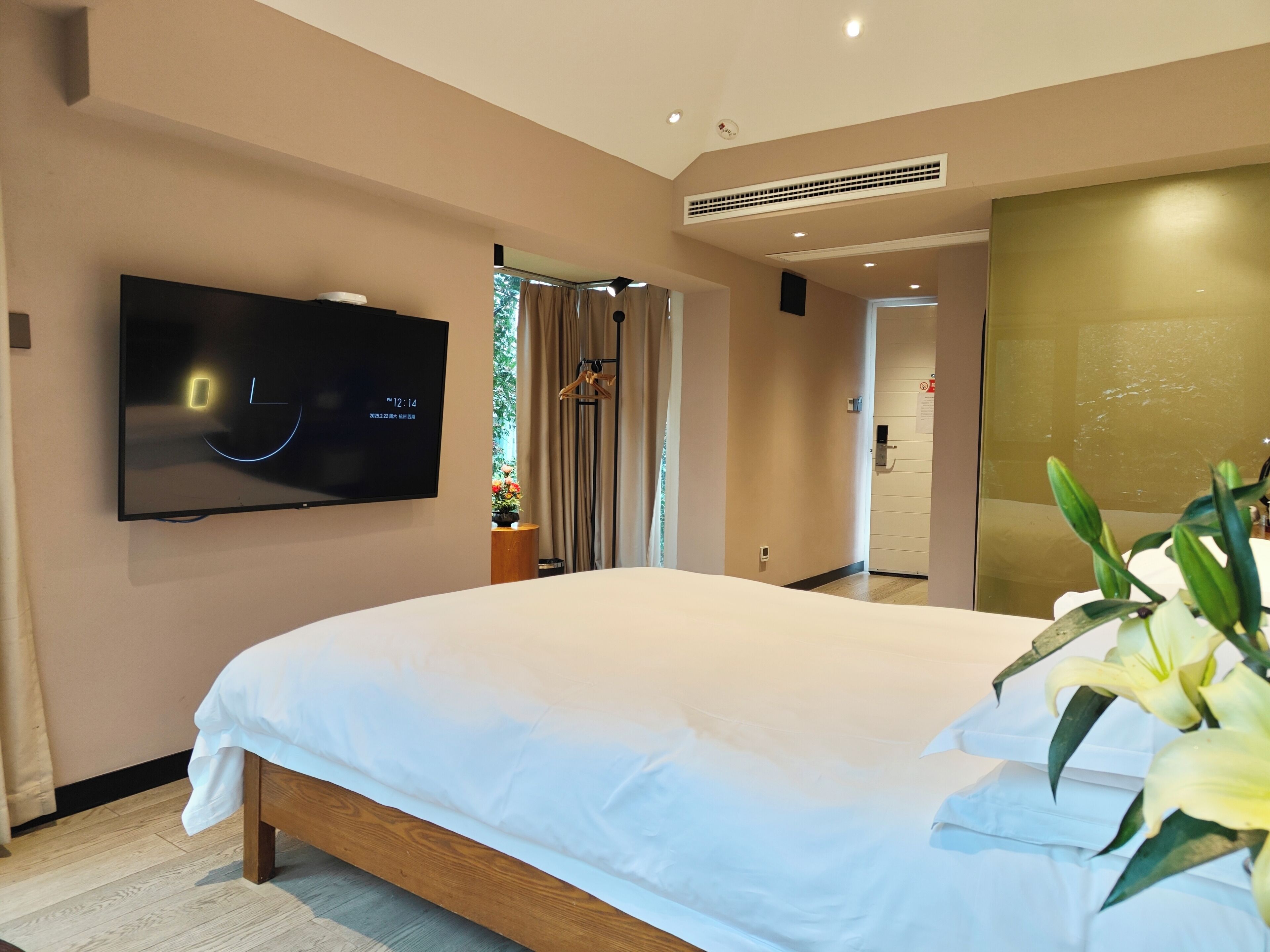 Una habitación de hotel moderna con una cama grande, un televisor de pantalla plana montado en la pared, una ventana con cortinas y una pequeña mesa con un jarrón de flores.