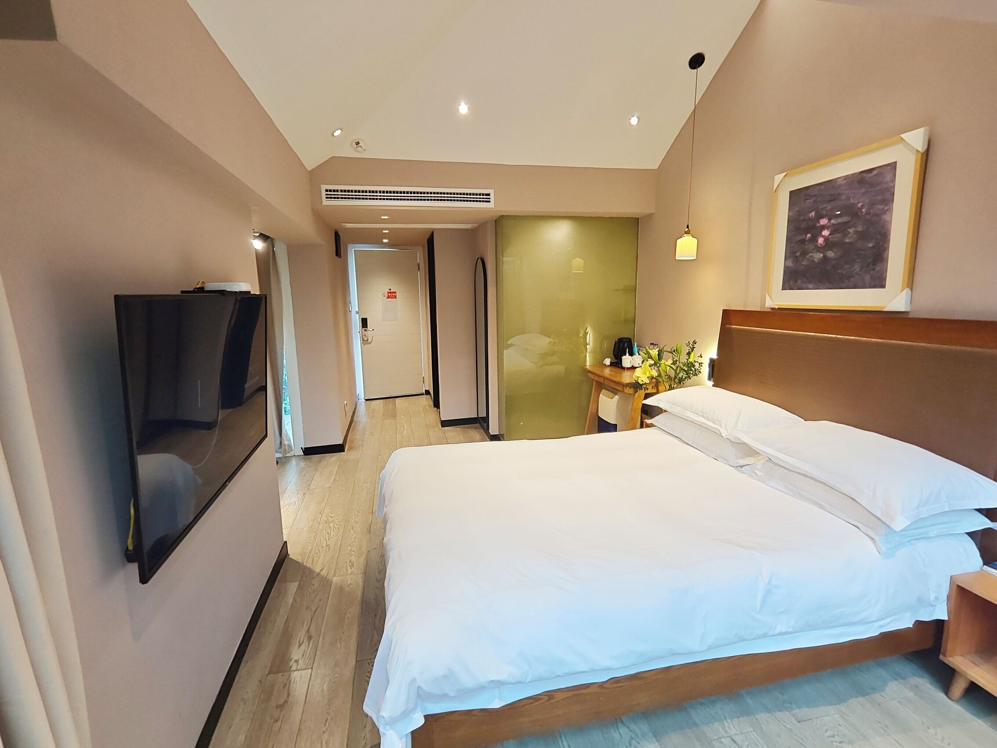 Una habitación de hotel con una cama grande, un televisor montado en la pared, una mesita de noche con una lámpara y un cuadro en la pared.