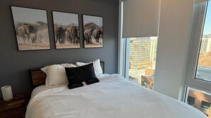 A bedroom with a large bed, a nightstand, a window with a view of buildings, and three framed pictures on the wall. - Modern 1BR Apt with Stunning Views and Pool Access (Toronto)