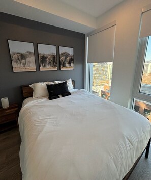 Ein Schlafzimmer mit einem großen Bett, einem Nachttisch, einem Fenster mit Blick auf Gebäude und drei gerahmten Bildern an der Wand.