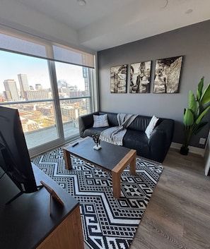 Ein modernes Wohnzimmer mit einem schwarzen Ledersofa, einem Holztisch und einem gemusterten Teppich. An der Wand hängen drei gerahmte Kunstwerke und ein großes Fenster mit Blick auf die Stadt.