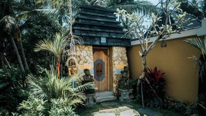 Una casa de piedra con puerta de madera, rodeada de plantas tropicales y un camino de piedras.