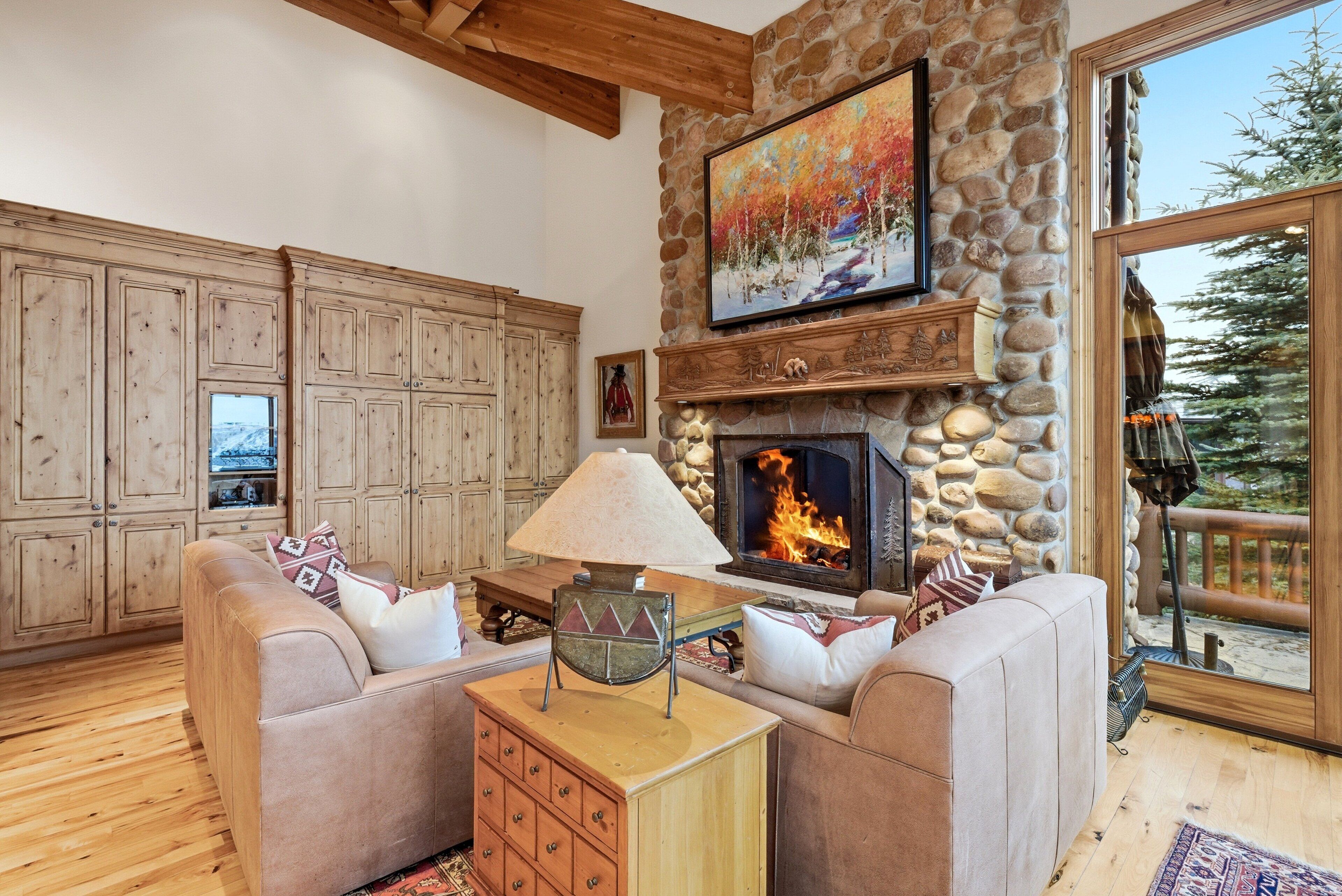 A cozy living room with a stone fireplace, wooden cabinets, and a large window overlooking a snowy landscape.