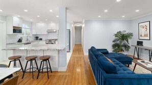 A modern kitchen with white cabinets, a marble island, and wooden flooring. There is a blue sofa and a plant in the living area. - Marbella Lane - Immaculate & Cozy 3BR Home (San Jose)
