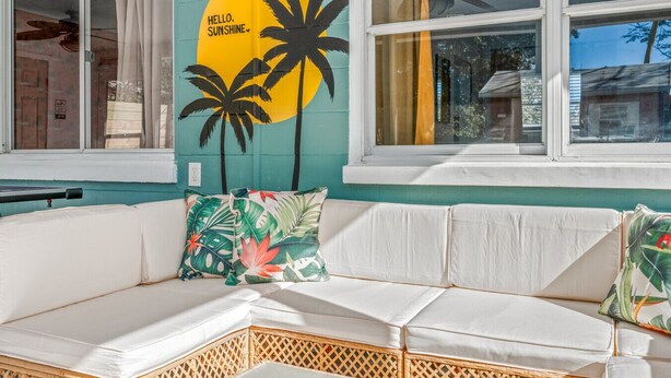 A white sectional sofa with tropical patterned cushions, a wall mural with palm trees and a sun, and a window with a view of the outside. - Beautiful East Orlando Home Near Everything (Orlando)